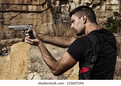 View of a menacing man pointing a handgun in a black shirt and dark shades on a stone quarry.
