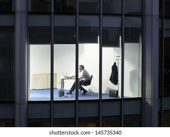 View Of Middle Aged Businessman Working Late Night At Office Desk Through Window