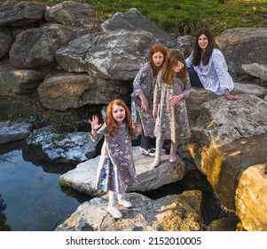 View Of Middle Aged Mother And Her Three Daughters Dressed In Similar Long Dresses Getting Over Landscaped Midwestern Stream; Youngest Girl Looking At Camera