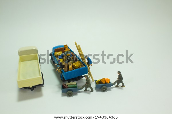 View of a miniature figure scene. A blue\
truck stands loaded with luggage while workers push luggage with\
one handcart to the other empty truck. White background. Concept\
loading and transportation
