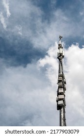 A View Of Mobile Communications Tower Situated In A Big Industrial City Against The Backdrop Of A Blue Sky, With White Fluffy Clouds. Cloudy Weather On A Background. No People Photography.