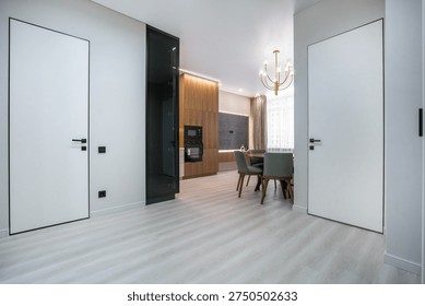 A view of a modern kitchen and dining space in an apartment. The room has light wood furniture and white walls. There are two doors visible on the sides. Natural light enters from a window