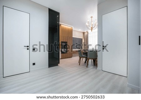 A view of a modern kitchen and dining space in an apartment. The room has light wood furniture and white walls. There are two doors visible on the sides. Natural light enters from a window
