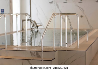View of modern pool with a metal handrails descent. Swimming, healthy and summer vacation concept. Steps into the pool with handrails in water, interior. The indoor pool rails with blue water. 