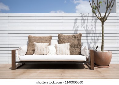 View of a modern simplistic white outdoor sofa on a deck of a house with a lone tree in a ceramic pot. Modern outdoor living