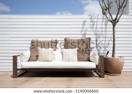 View of a modern simplistic white outdoor sofa on a deck of a house with a lone tree in a ceramic pot. Modern outdoor living