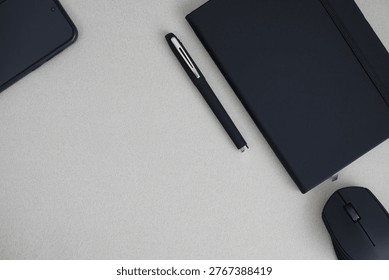 ​Top view of a modern tech workspace featuring a smartphone, black notebook, elegant pen, and wireless mouse on a grey textured surface with copy space.