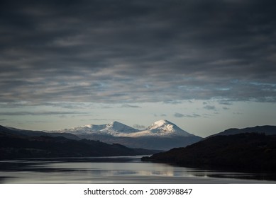 View of a mountain range covered in snow in Perth and Kinross, highlands of Scotland, United Kingdom, with snow-capped peaks reflected on the waters of the loch Tay under a dramatic sky
