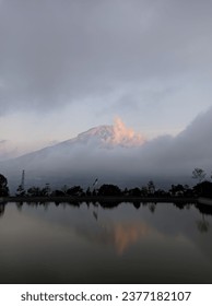 the view of mountains covered in clouds and trees that look black when illuminated by the setting sun makes a reflection at the bottom of the water