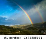 View of the mountains and a double rainbow. Part of the sky is open, part is occupied by a thundercloud.