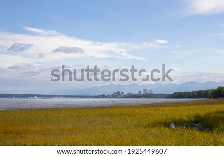 A view of the mud flats and downtown Anchorage, Alaska from the nearby coastal trail against a background of mountains and a cloudy sky; as seen in the summer of 2019.