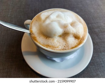 View Of A Mug Of A Cappuccino Coffee With Fluffy, White Milk Froth On The Table Covered With Neutral Grey Tablecloth In Front Of Window. Hot Drink In A Café