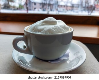 View Of A Mug Of A Cappuccino Coffee With Fluffy, White Milk Froth On The Table Covered With Neutral Grey Tablecloth In Front Of Window. Hot Drink In A Café