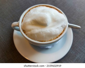 View Of A Mug Of A Cappuccino Coffee With Fluffy, White Milk Froth On The Table Covered With Neutral Grey Tablecloth In Front Of Window. Hot Drink In A Café