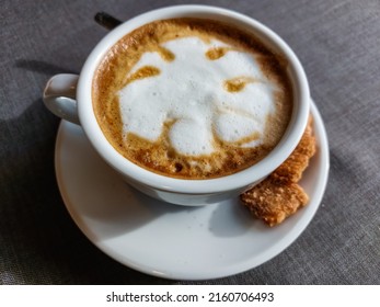 View Of A Mug Of A Cappuccino Coffee With Fluffy, White Milk Froth On The Table Covered With Neutral Grey Tablecloth In Front Of Window. Hot Drink In A Café