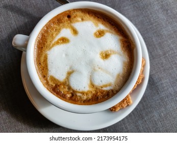 View Of A Mug Of A Cappuccino Coffee With Fluffy, White Milk Froth On The Table Covered With Neutral Grey Tablecloth In Front Of Window. Hot Drink In A Café