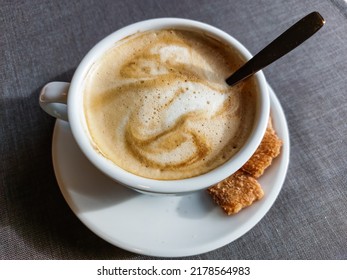 View Of A Mug Of A Cappuccino Coffee With Fluffy, White Milk Froth On The Table Covered With Neutral Grey Tablecloth In Front Of Window. Hot Drink In A Café