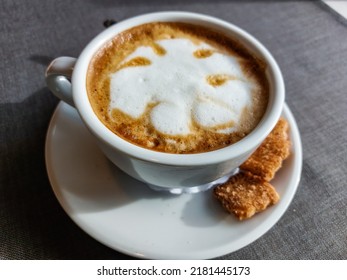 View Of A Mug Of A Cappuccino Coffee With Fluffy, White Milk Froth On The Table Covered With Neutral Grey Tablecloth In Front Of Window. Hot Drink In A Café