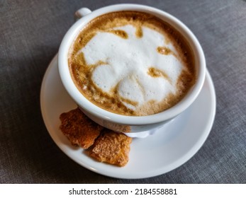 View Of A Mug Of A Cappuccino Coffee With Fluffy, White Milk Froth On The Table Covered With Neutral Grey Tablecloth In Front Of Window. Hot Drink In A Café