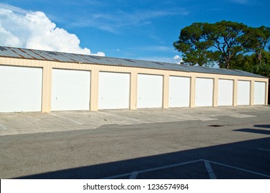 View Of Multi-unit Or Multi-bay Garage Type Storage Building Abandoned In Florida In Bright Sunshine With Large Tree And Clouds And Broken Driveway. Metal Roof Is Old, Damaged And Rusted.