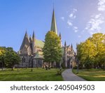 View of Nidaros Cathedral surrounded by lush green trees and set against a clear blue sky, with a well-maintained historic cemetery around it, bathed in the glow of a setting sun in Trondheim, Norway