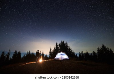 View of night starry sky over mountain valley with male hiker, illuminated camp tent and campfire. Man traveler standing near tourist tent under night sky with stars. Concept of night camping. - Powered by Shutterstock