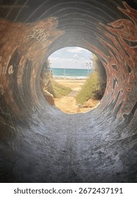 View of the ocean framed through a graffiti-covered tunnel. The sandy path outside leads to the turquoise sea, with waves rolling under a partly cloudy sky, creating a sense of escape and adventure.