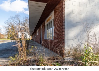 A View Of An Old Abandoned Strip Mall With Weeds Growing In Front Of Several Red Brick Store Fronts Closed After Long Struggle To Stay Open.