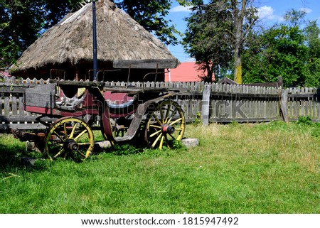 A view of an old devastated and damaged horse carriage or cart standing next to a wooden fence made out of planks or logs and a small wooden farmhouse with a thatched roof seen on a sunny summer day