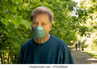 View Of Older Man Walking In Wooded Midwestern Park In Summer Wearing Protective Mask; Blurry Figures Of Couple With Dogs In Background