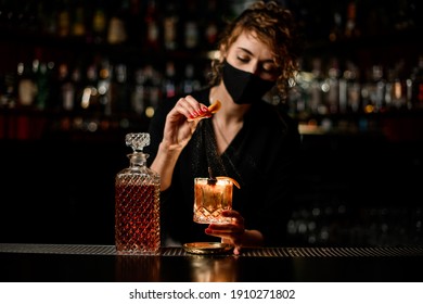 view on attractive woman bartender in mask who holds glass of cocktail and sprinkles on it