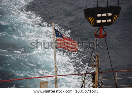 View on flying Liberian flag, stern light and secured hook of crane in aft station of cargo container vessel underway through calm ocean during summer sunny weather. Behind the vessel is keel water.