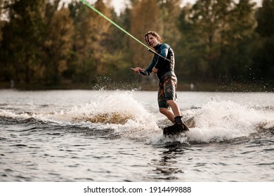 view on happy wet man in wetsuit balancing on wakeboard and rides down on the splashing wave on summer day