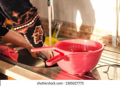 View On Kitchen Sink With Woman Hands Washing The Dishes With Water. A Pink Colander Is In Front. Cooking Process Concept.