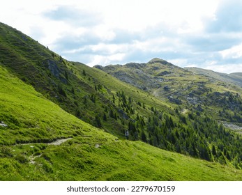 A view on lush green slopes of Austrian Alps in the region of Millstatt. There is a narrow pathway along the slope. Endless mountain chains in the back. Dense forest at the foothill. A bit of overcast
