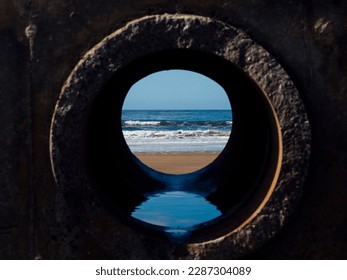 View on the ocean from a pipe or round shape hole with reflection inside the pipe. Abstract nature concept. Blue cool tone. Artificial framing of nature.