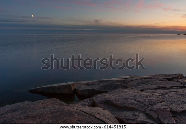 View on Onega Lake granite shore and\
evening glow with crescent reflecting in calm water at midnight\
sun. Besov Nos cape, Karelia Republic,\
Russia.\
