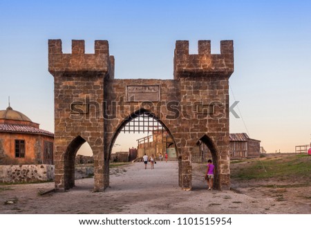 View on path through gates with towers with battlement and arches, house with dome in ancient fortress, all made of coarse stone, and walking people under clean blue sky just after sunset in Crimea