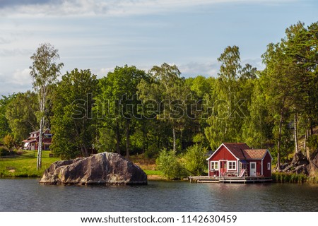 View on red holiday cabin by a lake in Stockholm archipelago, Sweden. Wooden cottage, sauna on shore. Tiny house near the water. Rocky small island, islet in water. Buildings surrounded by green trees