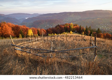 View on a wooden house in a mountain village. Wooden fence in the meadow with dry grass. Beautiful deciduous forest on the hill. Sunny weather. Carpathians, Ukraine, Europe