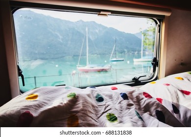 View out of a bus through a window with a caravan trailer with mountains in the background and the frame and bed in the foreground.                           
