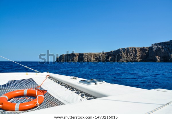 View out to sea on a white sailing boat with an\
orange life ring on the front netting. Blue sky and sea is divided\
by a dark rocky cliff\
face.