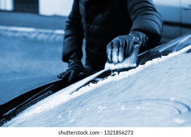 View From Outside The Car Of Woman Cleaning Car Windshield From Frost Using Specialized Ice Frost Scraper And Rubber Squeegee Car Windscreen In Cold Winter Blue Toned Image.