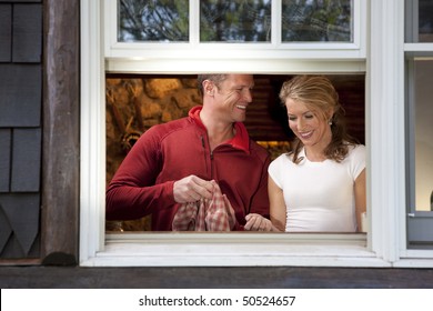 View From Outside A Window Of A Smiling Couple Doing Dishes Together In Their Kitchen. Horizontal Format.