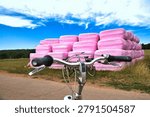 A view over a bicycle handlebar at a stack of hay bales wrapped in pink foil. A quirky rural scene by the roadside under a blue sky with clouds. A symbol against breast cancer.