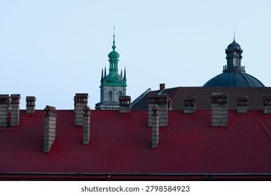 A view over red tiled rooftops and brick chimneys towards distant architectural landmarks against a clear sky