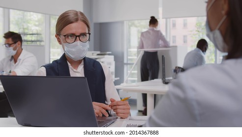 View Over Shoulder Of African Female Client In Safety Mask At Meeting With Bank Manager. Caucasian Real Estate Agent Woman In Protective Mask Consult Customer Sitting At Desk In Office