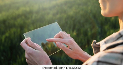 View over shoulder of female farmer in glass transparent screen in hands. Woman tapping on futuristic device while standing in green field. Touchscreen of hi-tech. Close up. - Powered by Shutterstock