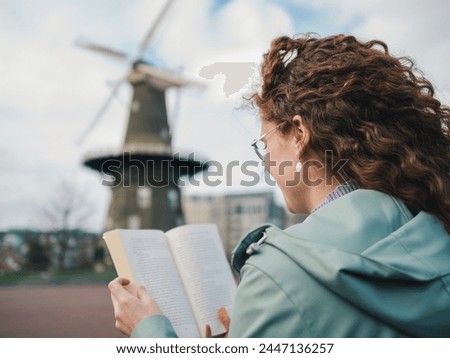 View over the shoulder of a woman with curly hair reading a book, with a traditional windmill in the blurred background