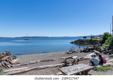 View Over A Small Beach With People Enjoying Some Lunch And Others Swimming On Tyler Street Plaza Park In Port Townsend, WA, USA With Views Over The Port Townsend Bay Against A Clear Blue Sky